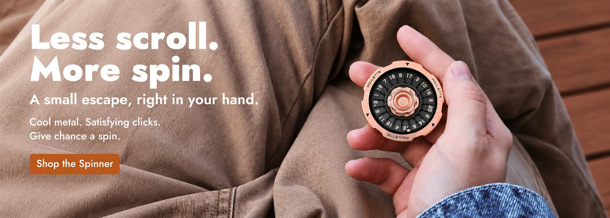 Eine Hand hält einen schwarz-roségoldfarbenen D20-Spinner über einer braunen Hose.