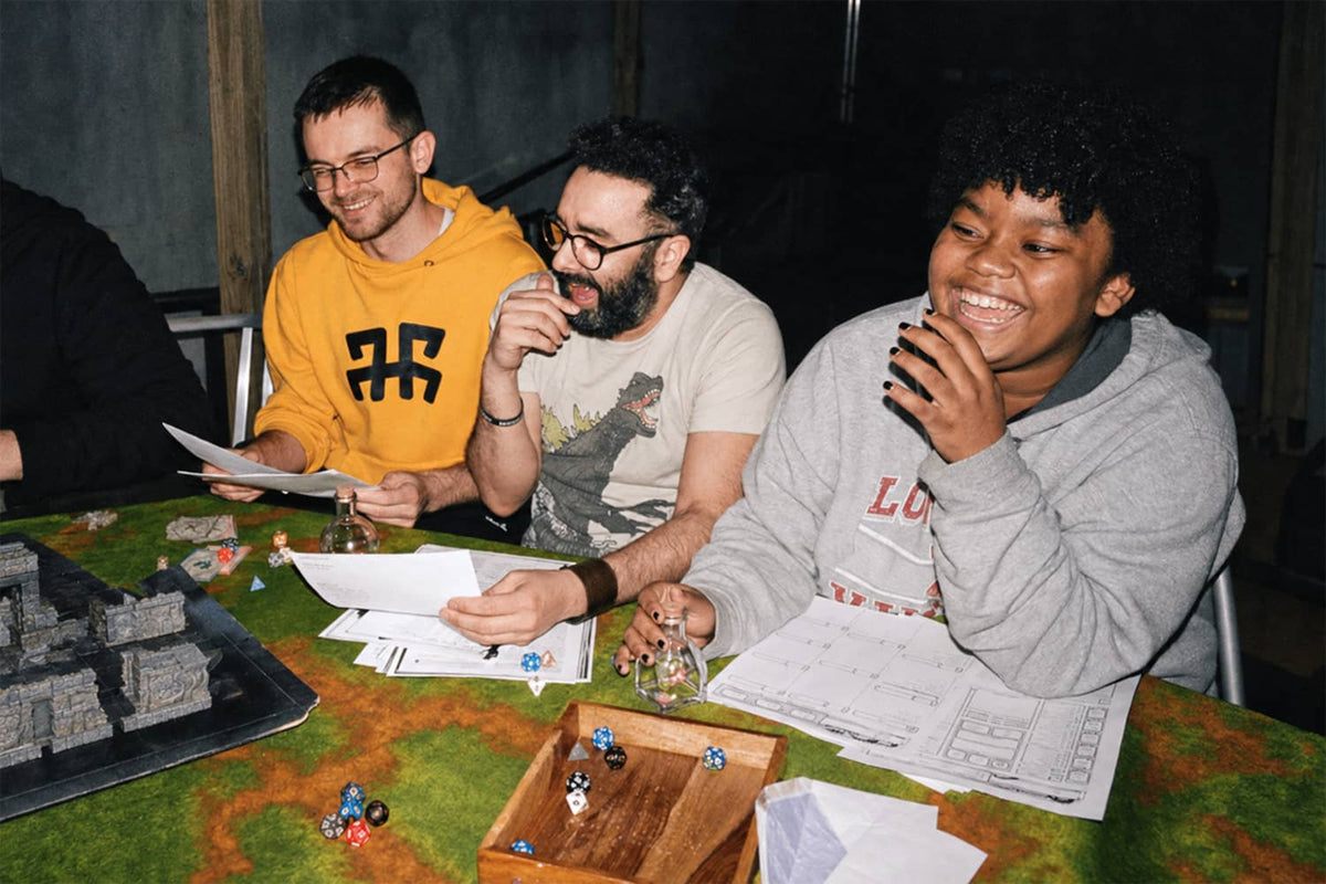 Drei Freunde lachen bei einem Rollenspiel am Tisch mit Würfeln und Karten.